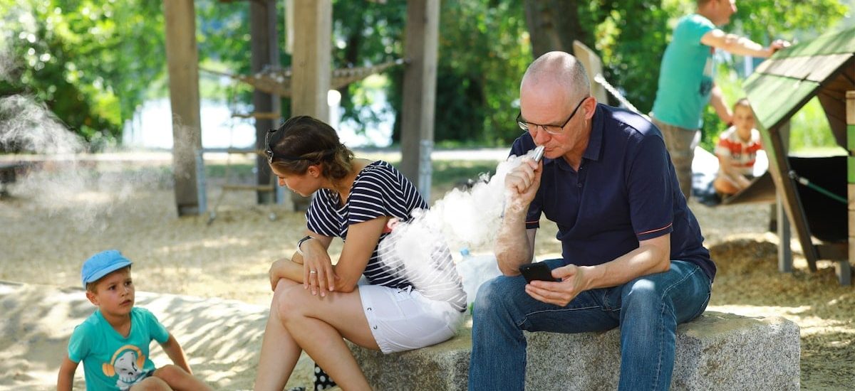 A men smokes a vape and disturbs a young mother and her son with his smoke – an appeal for more attention and awareness when smoking outdoors. Ein Mann raucht seine E-Zigarette auf einem Spielplatz und belästigt mit dem Rauch eine junge Mutter und ihren Sohn – ein Aufruf zu etwas mehr Rücksicht und Achtsamkeit beim Rauchen im Freien.